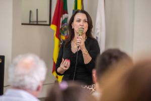 Foto horizontal que mostra uma mulher branca segurando um microfone. Ela tem cabelos castanhos longos e lisos, e veste uma blusa preta. Ao fundo, estão bandeiras do Brasil e do Rio Grande do Sul penduradas em hastes de madeira, que se destacam sobre uma parede branca. À frente, aparecem cabeças de pessoas sentadas, levemente desfocadas.