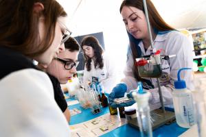 Foto horizontal. À esquerda, um menino e uma menina (ambos de óculos), observam uma mulher de cabelos longos, castanhos e lisos (vestida de jaleco branco e luvas azuis) manipular objetos sobre uma mesa, repleta de tubos de ensaio, pivetas e outros recipientes tipicamente usados em laboratórios. Ao fundo da foto, há uma outra jovem de cabelos longos e castanhos, usando óculos e também vestida com jaleco branco