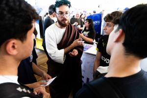 Foto horizontal. Ao centro, um rapaz de barba e óculos veste uma fantasia que remete à Grécia ou Roma antigas, incluindo um objeto verde representando louros em volta da cabeça. Ao redor dele, estão quatro adolescentes (meninos vestidos de preto).