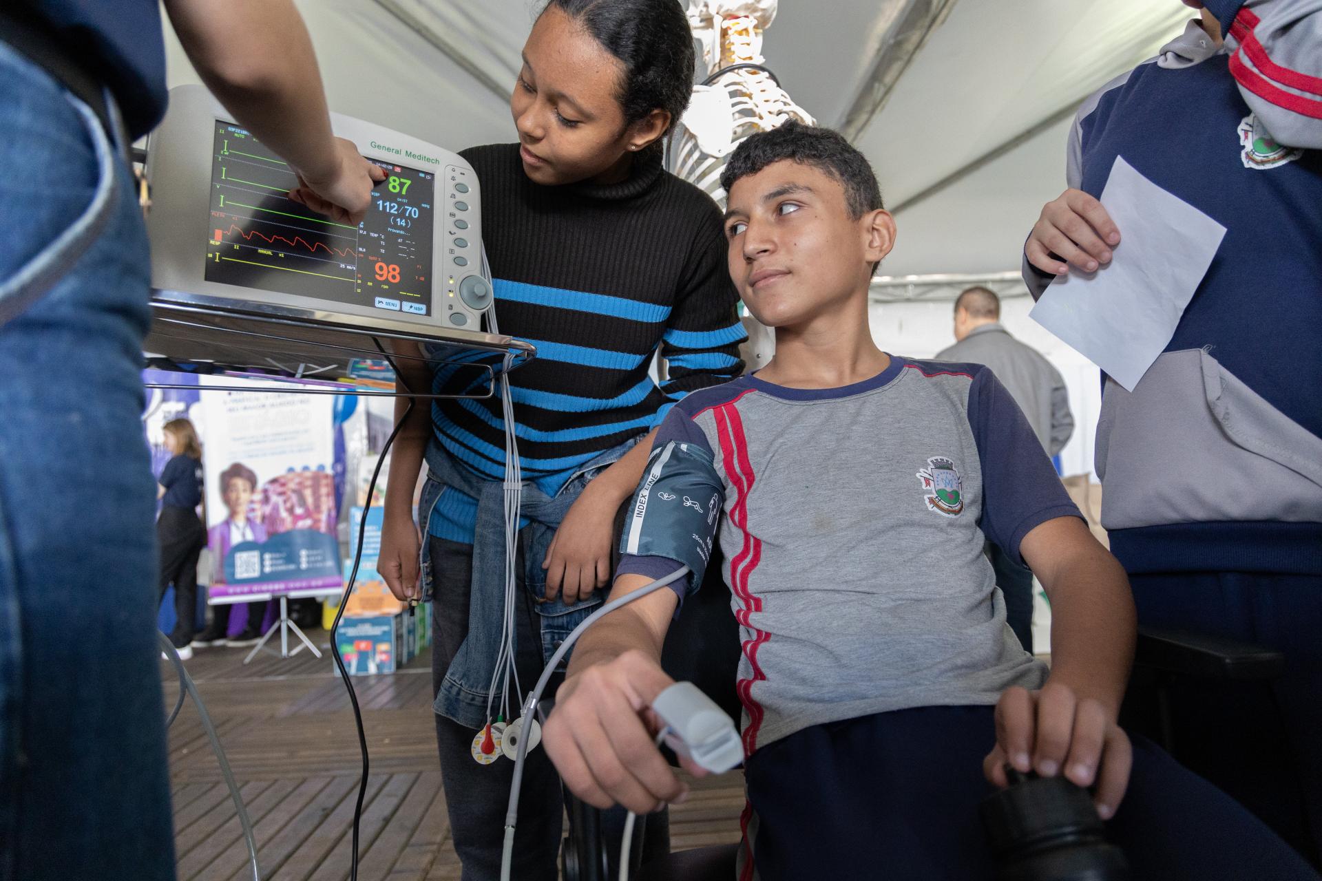 Foto horizontal: A imagem foca em quatro pessoas, das quais se pode ver o rosto de apenas duas – ambos de pele morena e cabelo escuro, vestindo calças pretas. A menina, em pé, tem cabelo comprido e veste uma blusa preta com listras horizontais azuis. O menino, sentado, veste uma camisa cinza com detalhes em azul e vermelho, com o brasão de Santa Maria no lado esquerdo do peito, o que dá a entender que é aluno de uma escola municipal. No seu braço direito, lhe foram acoplados dispositivos para medir pressão e temperatura. Ele olha à sua direita para o aparelho ao qual os dispositivos estão conectados. Uma pessoa, no lado esquerdo da foto (fotografada da cintura para baixo), vestindo calças jeans, aponta um dedo da mão direita para o visor do aparelho.