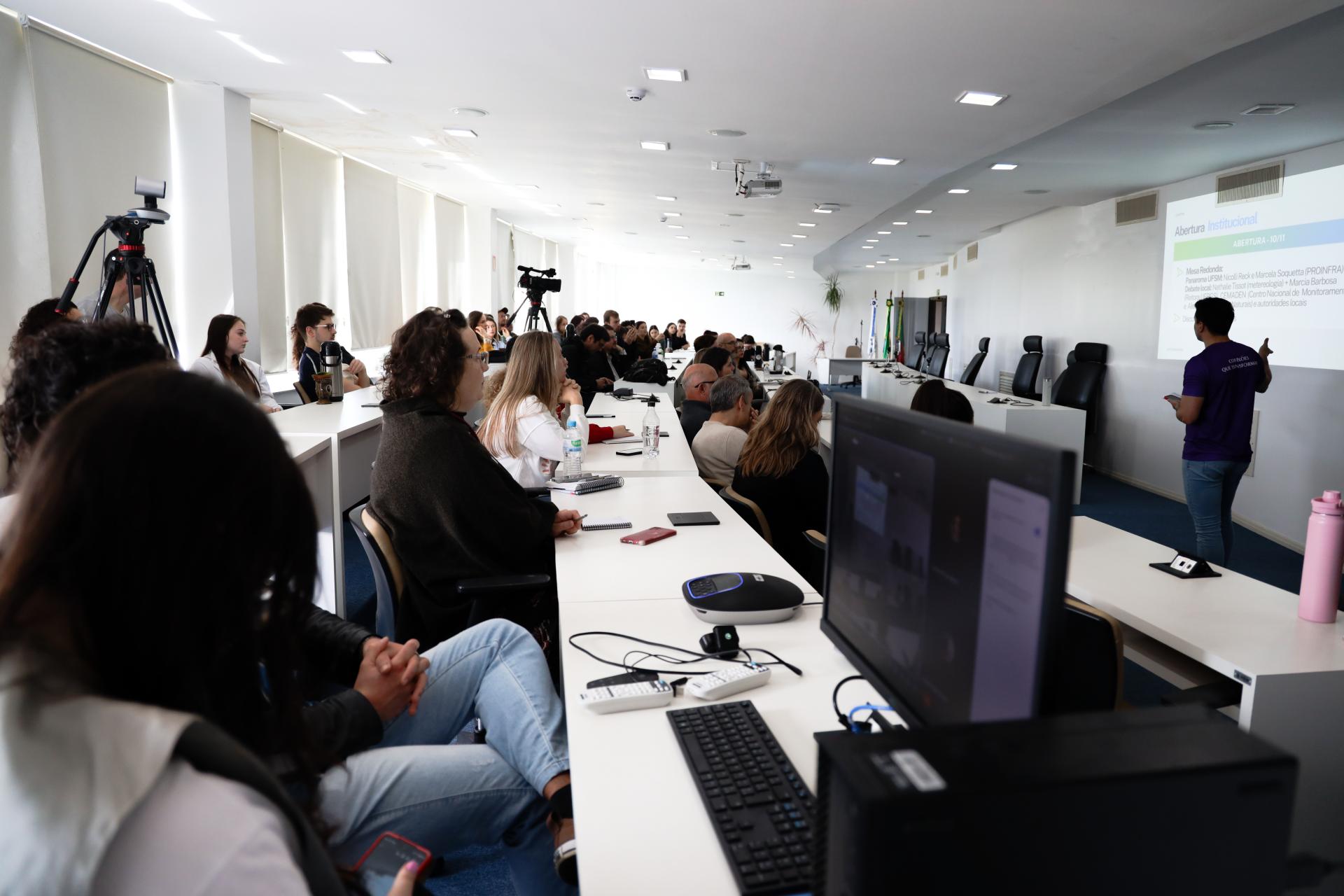 Foto horizontal colorida, que retrata uma apresentação em uma sala para um grupo de pessoas. À esquerda e ocupando quase todo espaço da cena, pessoas estão sentadas em fileiras de cadeiras, algumas anotando em cadernos ou segurando celulares. À direita, um homem de camiseta roxa com a inscrição “Conexões que transformam” está em pé apresentando, diante de uma parede branca com projeção de texto em tela.