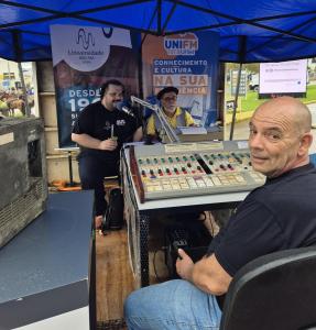 Foto horizontal. Embaixo de uma lona azul e com cartazes das rádios da UFSM ao fundo, três homens estão em volta de uma mesa de áudio. Um deles, à esquerda, fala ao microfone