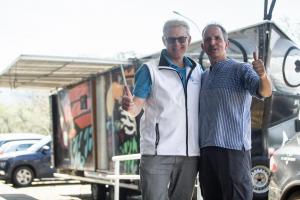 Fotografia colorida e horizontal que mostra dois homens fazendo sinal de positivo para a câmera. O homem à esquerda utiliza uma camisa azul e uma calça preta, enquanto o homem à direita utiliza um colete branco sobre uma camisa azul e calça cinza. Ao fundo é possível ver o trailer, que na sua parte frontal conta com olhos desenhados e antenas decorativas representando uma borboleta. A cor predominante da carroceria do trailer é preta e, sobre ela, há detalhes grafitados nas cores vermelha, verde, azul, laranja e branca.