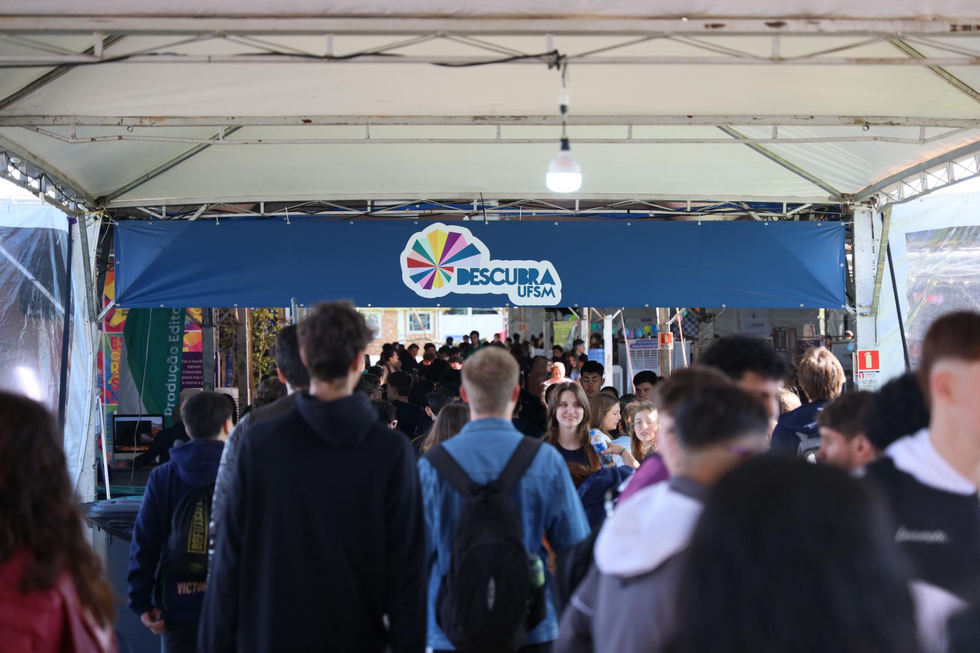 Foto colorida horizontal do Descubra. A imagem mostra um local com fechado com uma estrutrua de lonas e com muitas pessoas com roupas casuais em pé. Ao fundo, um toldo em azul com a marca do Descubra.