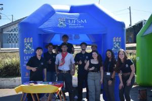Foto horizontal. Mostra alunos enfileirados posando para foto, sob uma barraca inflável da UFSM, de cor azul.