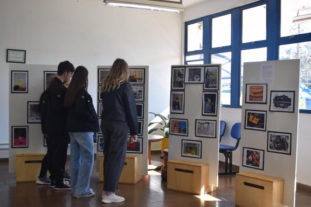 Foto colorida horizontal. Quatro pessoas jovens observam mostra. Dos quatro jovens, três são meninas e um é menino. Todos usam roupas casuais, como jaqueta jeans. À frente deles, quadros retangulares com várias fotografias de diferentes estilos, coloridas e em preto e branco.