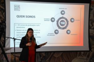 Foto horizontal e colorida de Débora Flores, mulher branca vestindo roupa social nas cores preta e laranja, apresentando um slide do Hub.Doc