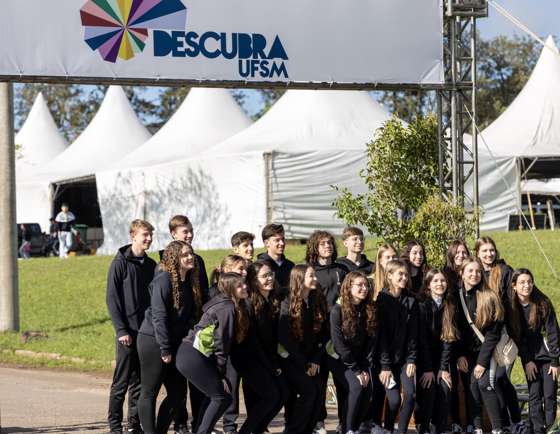 Foto horizontal e colorida de um grupo pequeno de estudantes vestindo roupas casuais nas cores pretas. Eles posam em grupo para uma fotografia em frente ao pórtico de entrada do Descubra 2025