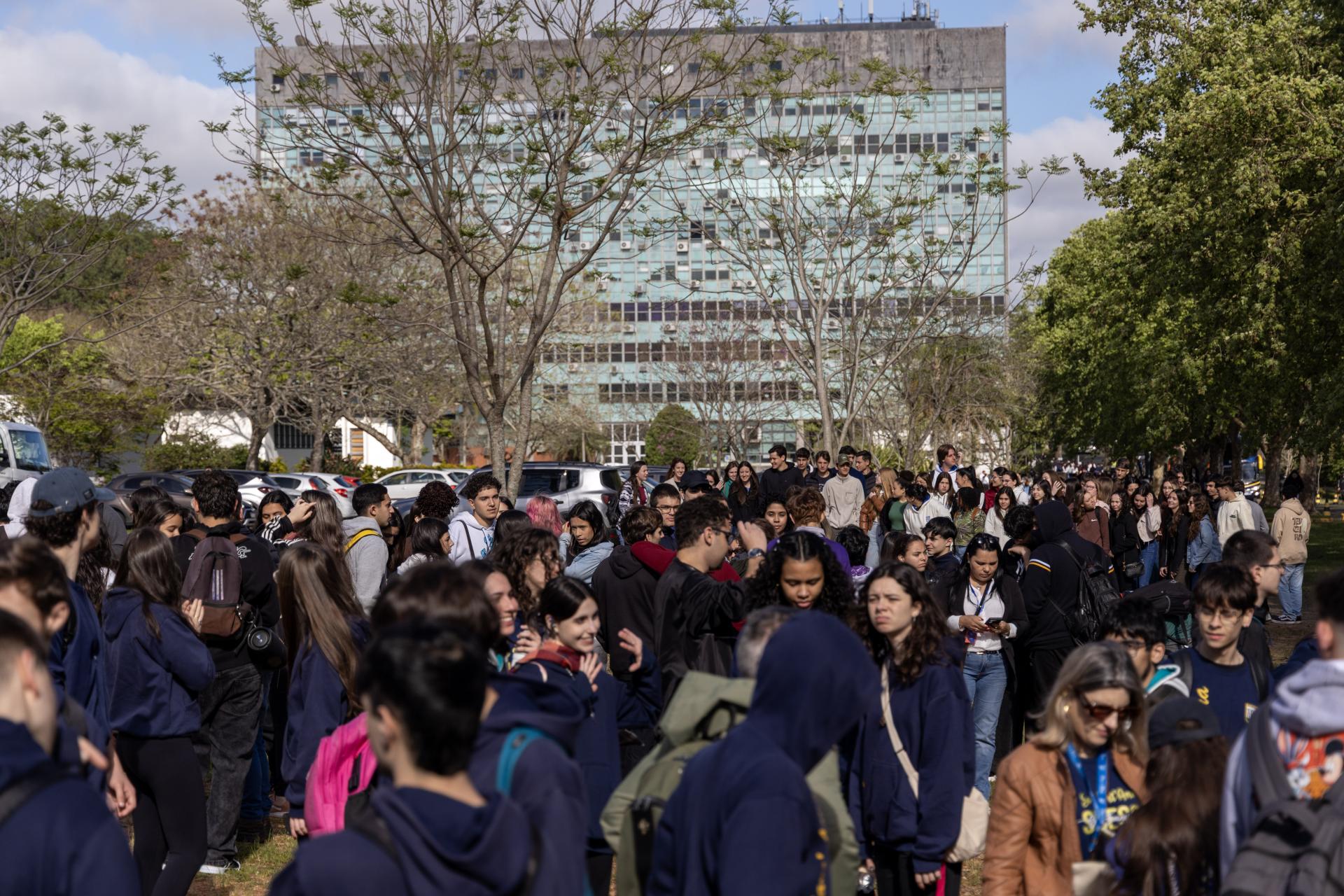 Foto horizontal e colorida de um grande de estudantes vestindo uniforme em cores predominantemente azuis marinhos. Ao fundo, está o prédio da Reitoria da UFSM