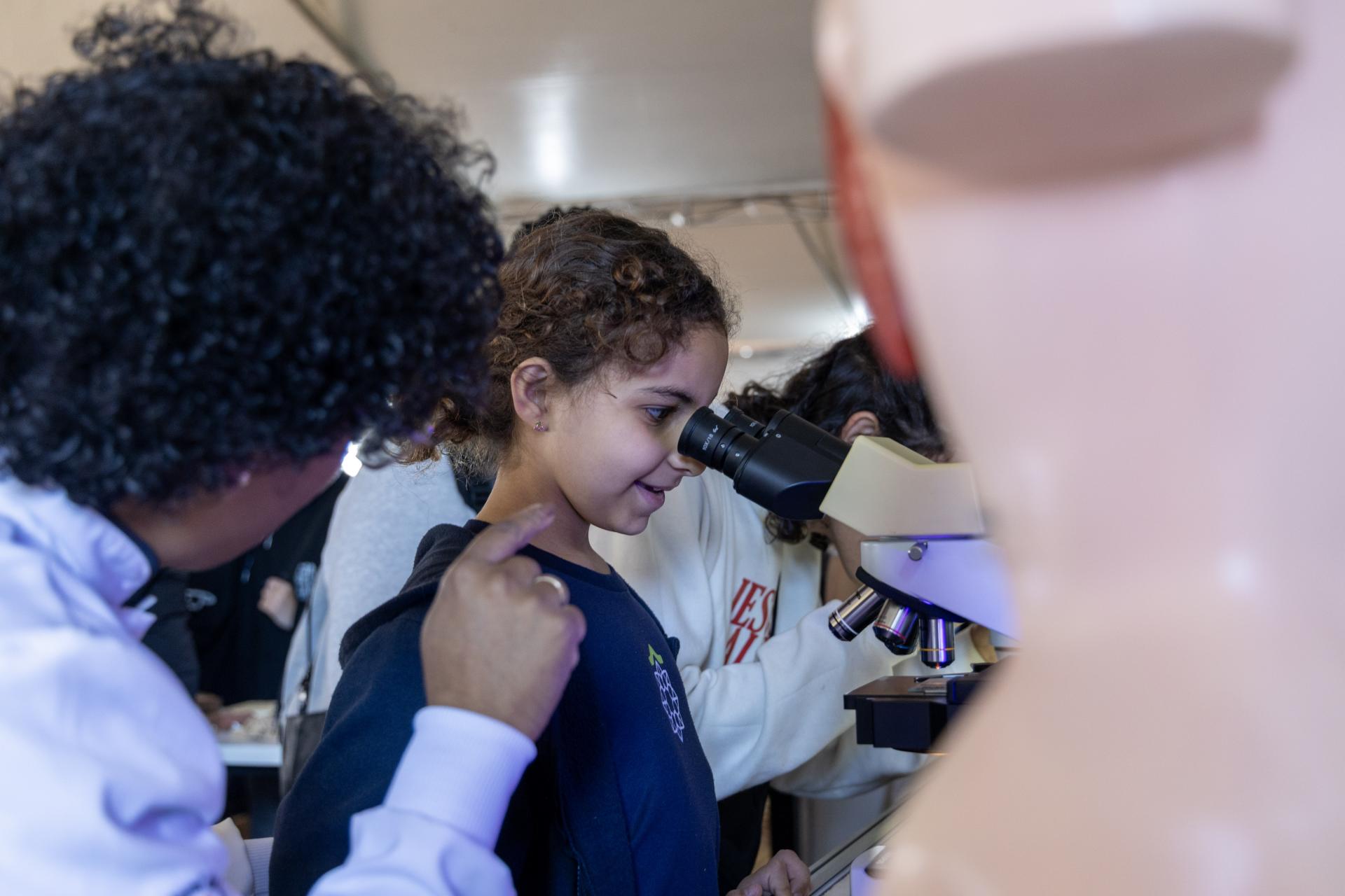 Foto horizontal e colorida de uma estudante criança observando um aparelho microscópico. Ao lado esquerdo da foto, uma mulher aparece observando a aluna