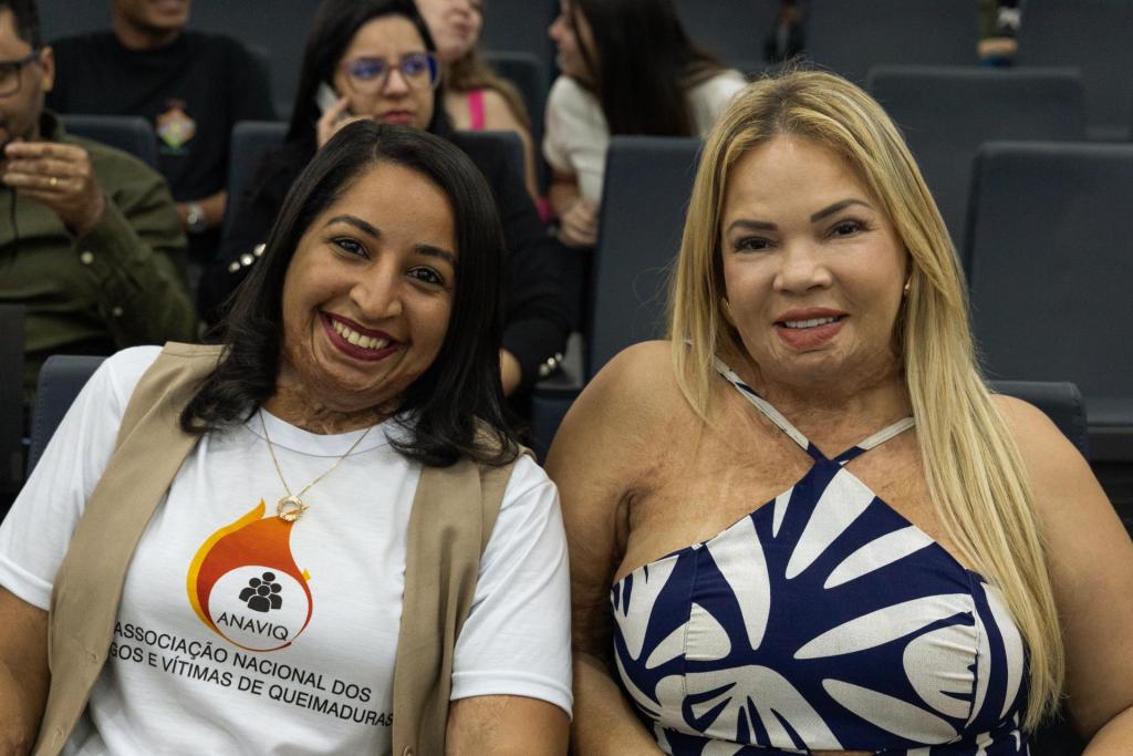 Fotografia horizontal. Mostra duas mulheres que sorriem. À esquerda Adrielle Macedo, uma mulher com cabelos pretos, curtos e lisos. Ela veste uma blusa que diz “Associação Nacional dos Amigos e Vítimas de Queimaduras”. Ela olha para a câmera. À direita está Rhafrezzya Alves, uma mulher loira, com cabelos lisos e longos. Ela usa um vestido azul e branco longo, com alças finas.