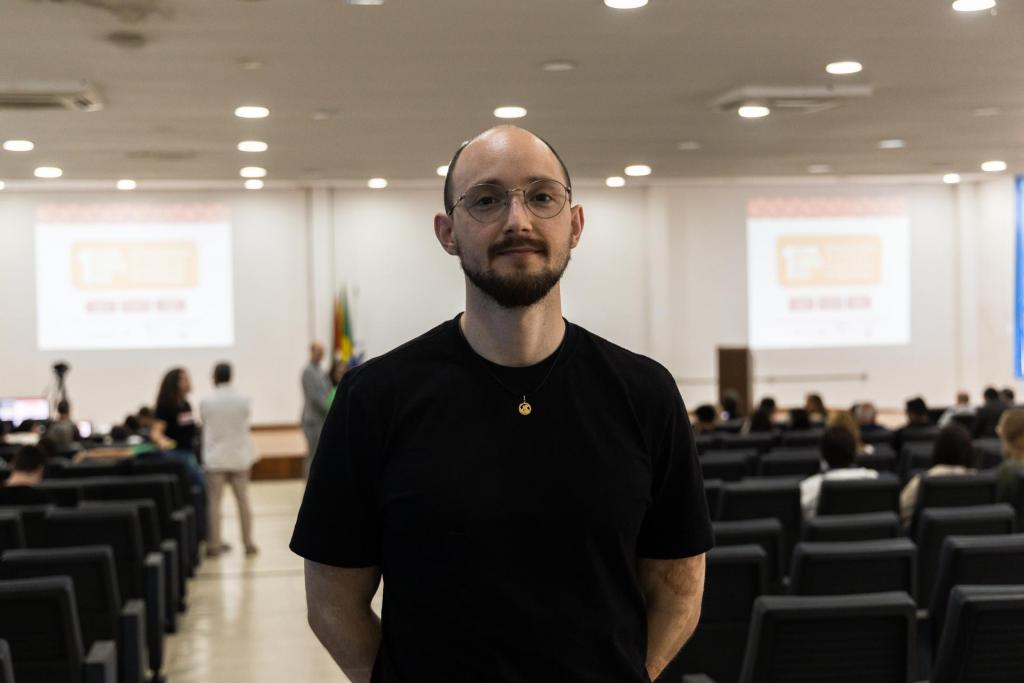 Fotografia horizontal. Mostra um homem que olha para a câmera. É Delvani Rosso, um homem alto, calvo, com óculos com armação redonda. Ele veste uma camisa preta lisa. Ao fundo está a sala onde o congresso.