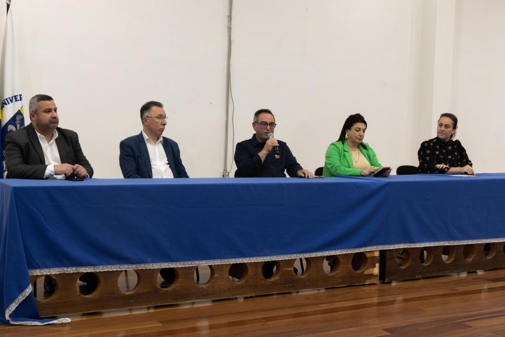 Fotografia horizontal, Mostra uma mesa grande com cinco pessoas. Mais à esquerda está Edson Roberto, um homem de cabelos pretos e curtos, que veste roupa social. Ao lado direito dele está Valdeci Oliveira, vestindo um paletó azul, uma camisa branca e uma calça de alfaiataria da mesma cor do paletó. À direita de Valdeci está Flávio José da Silva. Ele veste uma camisa preta e calça jeans escura. À sua direita está Martha Adaime que veste um blazer verde limão e uma blusa bege. Ao lado direito de Martha está Marina Callegaro, que está com o cabelo preso e veste uma blusa preta.