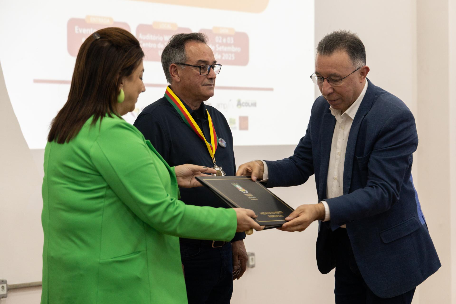 Fotografia horizontal. Ao meio está Flávio José da Silva. Ele veste uma camisa preta e calça jeans escura. Em seu rosto utiliza um óculos com armação quadrada. O destaque vai para o item em seu pescoço, a Medalha do Mérito Farroupilha. Mais à esquerda está Martha Adaime, com seu corpo virado para a direita, ela veste um blazer verde limão e segura o certificado da Medalha do Mérito Farroupilha. Mais à direita está Valdeci Oliveira, vestindo um paletó azul e óculos quadrados. Ele está virado para a esquerda, junto de Martha, segura o certificado.