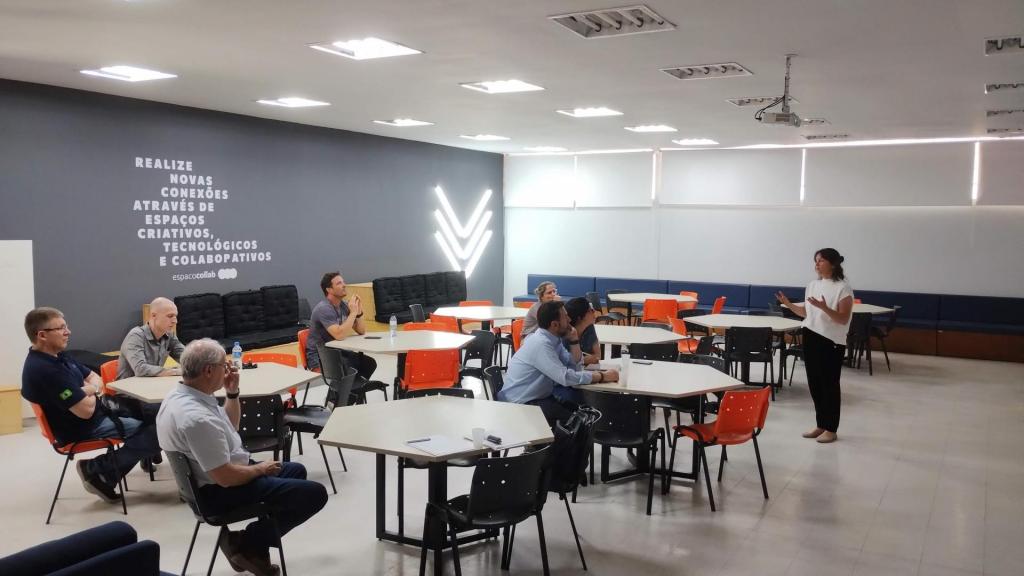 Foto colorida horizontal. A imagem mostra, em uma sala de aula, do lado direito, a tradutora Amy em pé, à frente de um grupo de sete pessoas sentadas, sendo cinco homens e duas mulheres. Amy usa roupa casual e tem as mãos inclinadas. O grupo está sentado de forma distribuída em cadeiras laranjas ao redor de mesas hexagonais. Ao fundo há duas paredes, uma branca lisa, e outra preta, com palavras brancas escritas nela: “Realize novas conexões em espaços criativos, tecnológicos e criativos”.