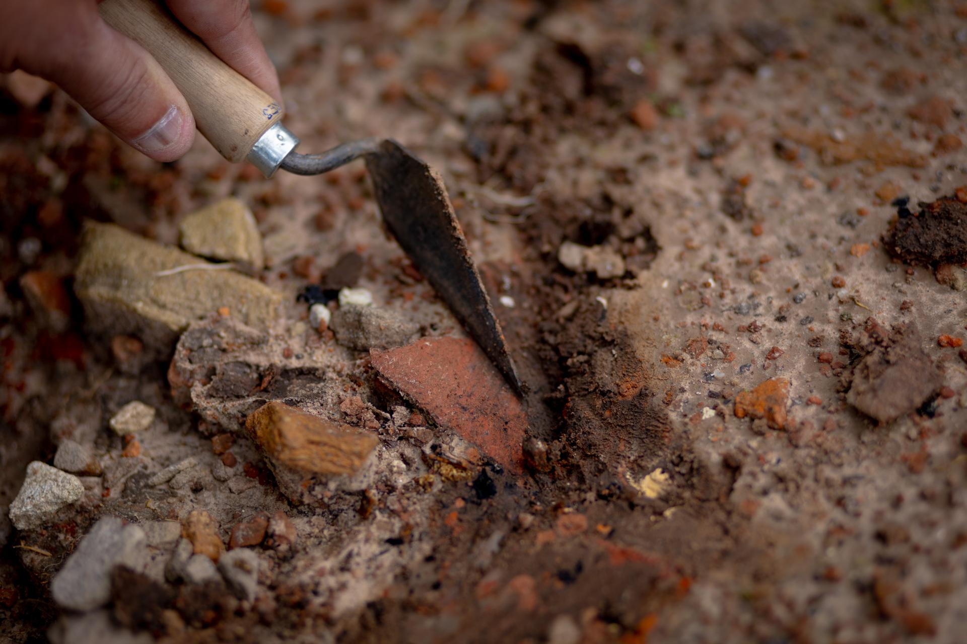 Foto colorida horizontal de mão escavando terra com uma ferramenta, que tira a terra de cima de um pedaço de telha.