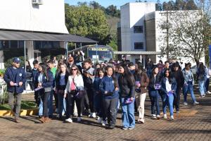A imagem mostra um grupo grande de pessoas caminhando em área externa, no campus da UFSM em Frederico Westphalen. Na frente, há homens e mulheres adultos conduzindo jovens estudantes. Muitos carregam folhetos de cor roxa. o fundo, aparece um ônibus escolar amarelo. Há árvores e vegetação ao redor, e o clima é de dia ensolarado.