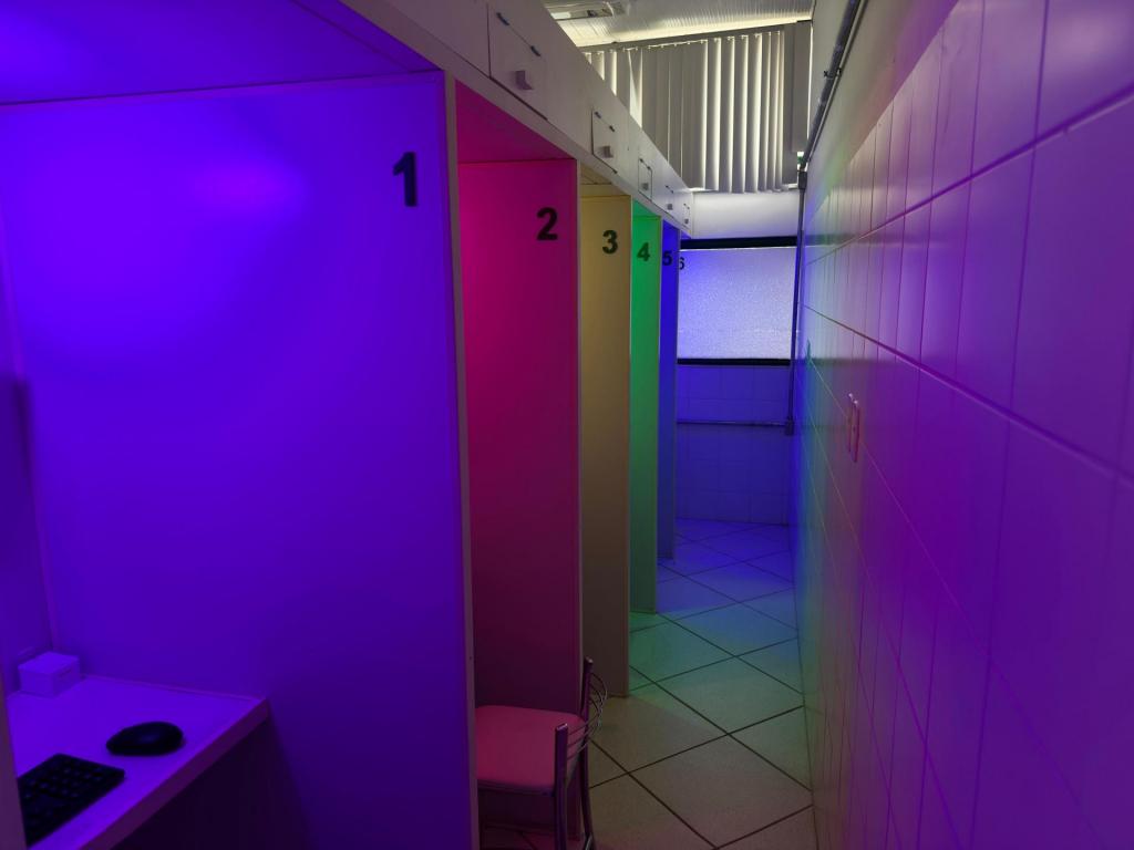 Fotografia horizontal colorida. A esqueda cabines numeradas de um a cinco, cada uma com uma luz de uma cor diferente ligada, respectivamente: lilás, vermelho, amarelo, verde e azul. Ao centro um corredor que leva até uma janela no fundo da sala. À direita uma parede de azulejos brancos que reflete as cores da iluminação.