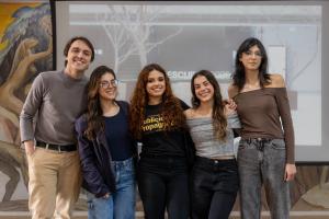 Foto colorida, horizontal. Cinco pessoas posam sorrindo em pé, com roupas casuais, em frente a uma projeção com o texto “Descubra 2025”.