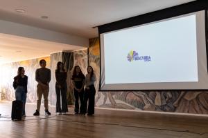 Foto colorida, horizontal. Cinco pessoas com roupas casuais estão em pé lado a lado, voltadas para a direita, próximas a uma tela de projeção com o logo “Descubra UFSM”.