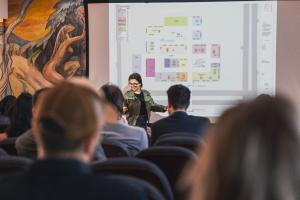 Foto colorida, horizontal. Uma mulher em pé, sorridente e de óculos, apresenta diante de uma tela com planta baixa projetada e legenda colorida.