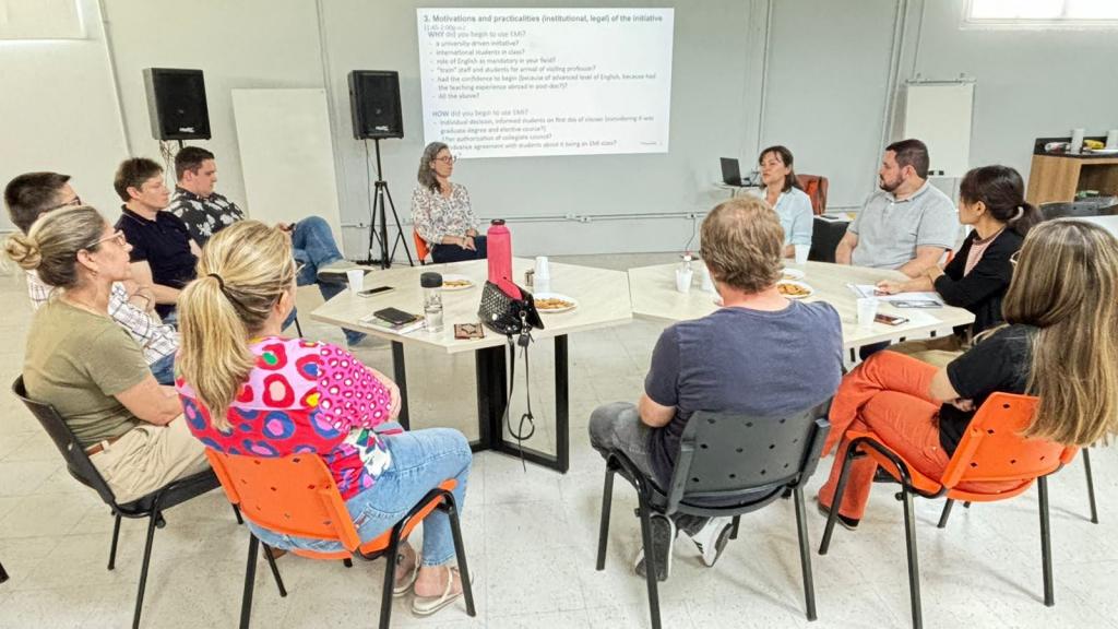 Foto colorida horizontal. Um grupo de onze pessoas, incluindo a tradutora Amy, usa roupas casuais e está em formato de círculo ao redor de duas mesas. As mesas tem formato de hexágono, e em cima dela há celulares, garrafas, papéis, uma pequena bolsa e pratos com comida. Ao fundo há caixas de som e uma imagem projetada na parede, com um texto