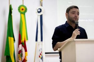 Um homem segurando um microfone está falando. É um homem branco, de cabelo castanho curto e barba, usando uma camisa azul escura. Ele está atrás de um palanque, e do lado esquerdo ao fundo há três bandeiras, da esquerda para a direita: do Brasil, do Rio Grande do Sul, e da Universidade Federal de Santa Maria.