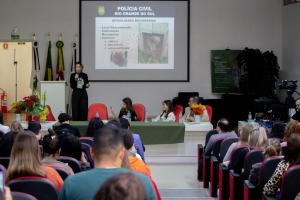 Um grupo de pessoas está em um auditório assistindo a uma palestra. Na parte inferior da foto, estão as pessoas assistindo, sentadas em cadeiras vermelhas e pretas. Na parte central da foto, há uma mesa com um pano verde no centro e vasos com flores nos dois cantos. Três pessoas estão sentadas, e uma mulher está de pé. A mulher é branca, tem cabelo curto, loiro escuro e ondulado, usa roupas pretas e um crachá verde de papel. Ela segura um microfone e está falando. Ao fundo, há um banner do evento no canto direito, no centro há uma imagem projetada com slides, no slide está escrito Polícia Civil do Rio Grande do Sul, e no canto esquerdo há várias bandeiras, um palanque, uma porta e um extintor de incêndio.