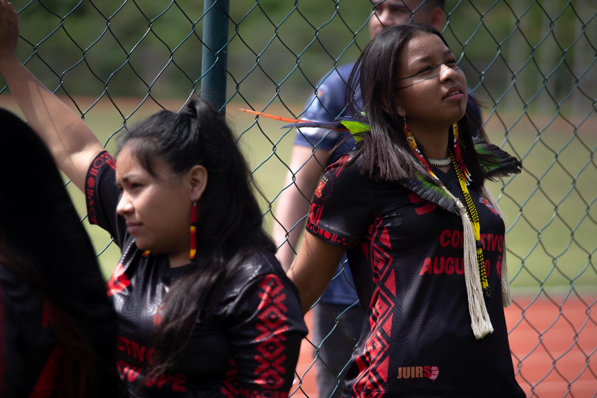 foto colorida horizontal de duas jovens indígenas, usando brincos e adereços com penas, em um espaço ao ar livre