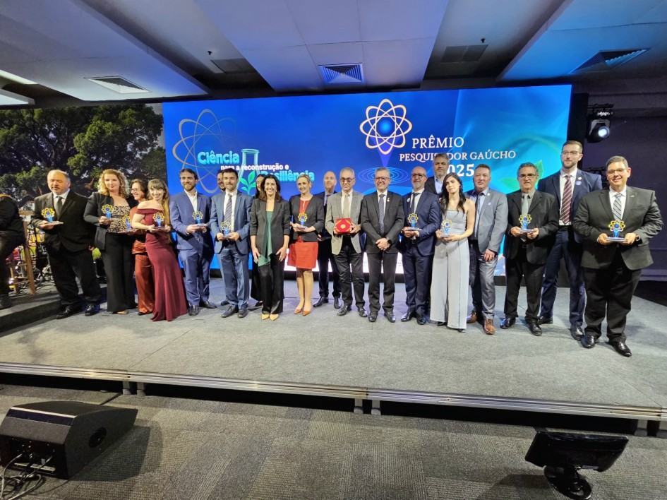 Fotografia colorida com pessoas alinhadas lado a lado, segurando os prêmios, em cima de um palco