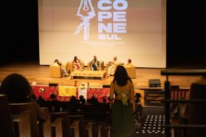 Foto horizontal. Mostra um auditório com filas de cadeiras em forma de degraus, semelhante a um cinema. Ao centro, uma mulher de costas (de cabelo escuro, vestindo blusa e saia verdes) desce uma das escadas da plateia. No palco, tendo ao fundo a logo marca do Copene, cinco pessoas estão sentadas em poltronas em torno de uma mesa.