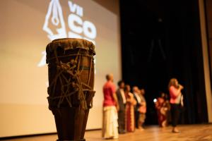 Foto horizontal. Mostra em primeiro plano, à esquerda, um tambor de madeira de cerca de um metro de altura, enrolado em cordas. À direita e ao fundo, estão oito pessoas em pé (todas elas desfocadas). Uma mulher à frente do grupo fala ao microfone.