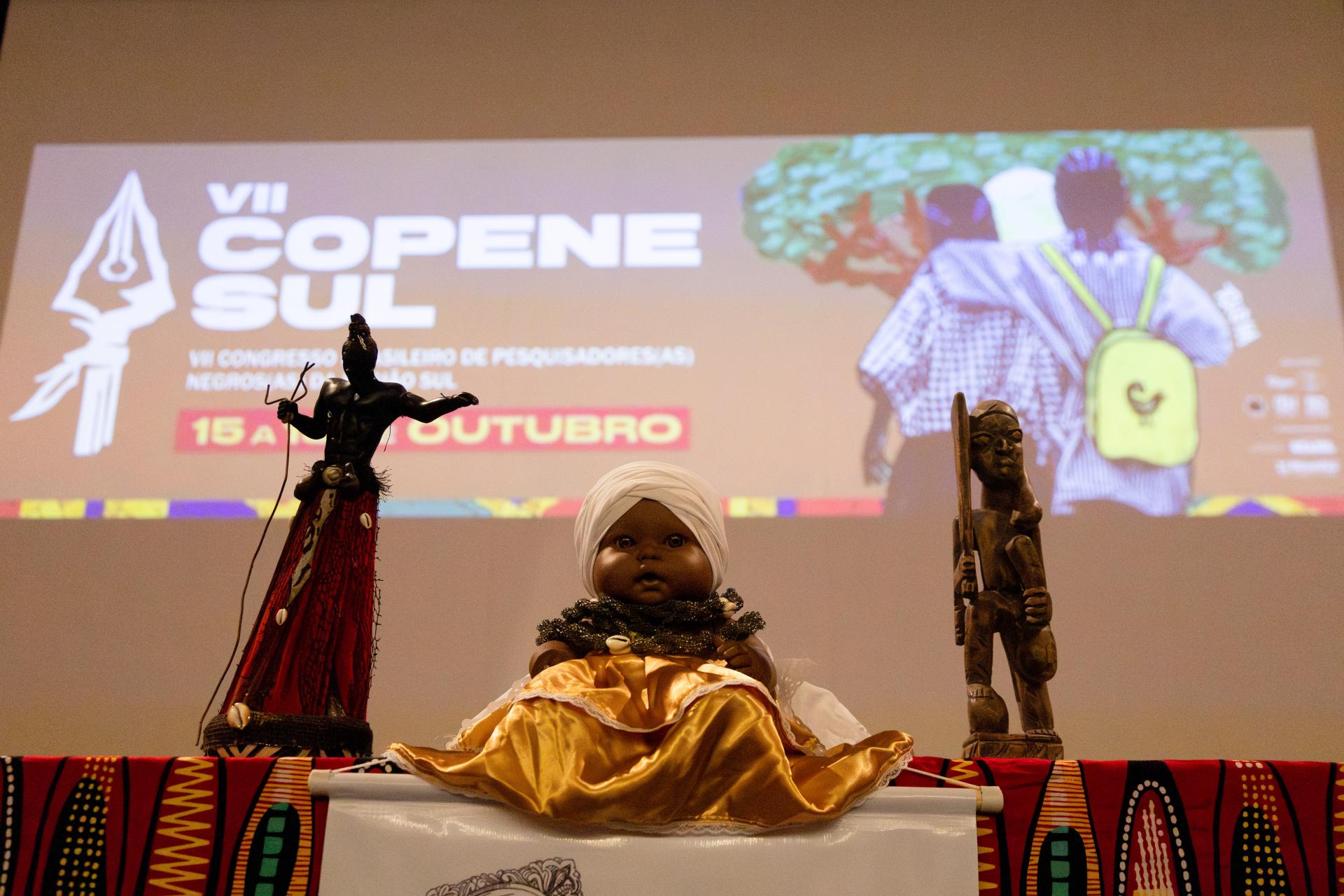 foto colorida horizontal de duas estátuas de negros e uma cabeça de boneca negra dispostos como objetos decorativos no palco onde ao fundo está projetado o banner do evento
