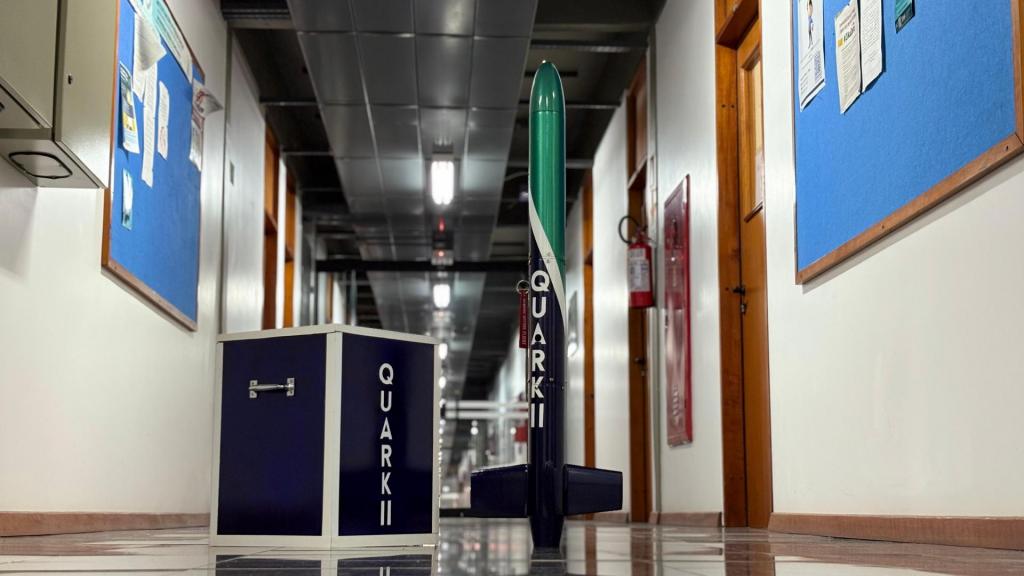 Foto colorida horizontal de foguete preto, verde e branco ao lado de um cubo preto com inscrições em branco. O foguete tem mais de um metro e meio e está no situado no chão em um corredor de um prédio da universidade