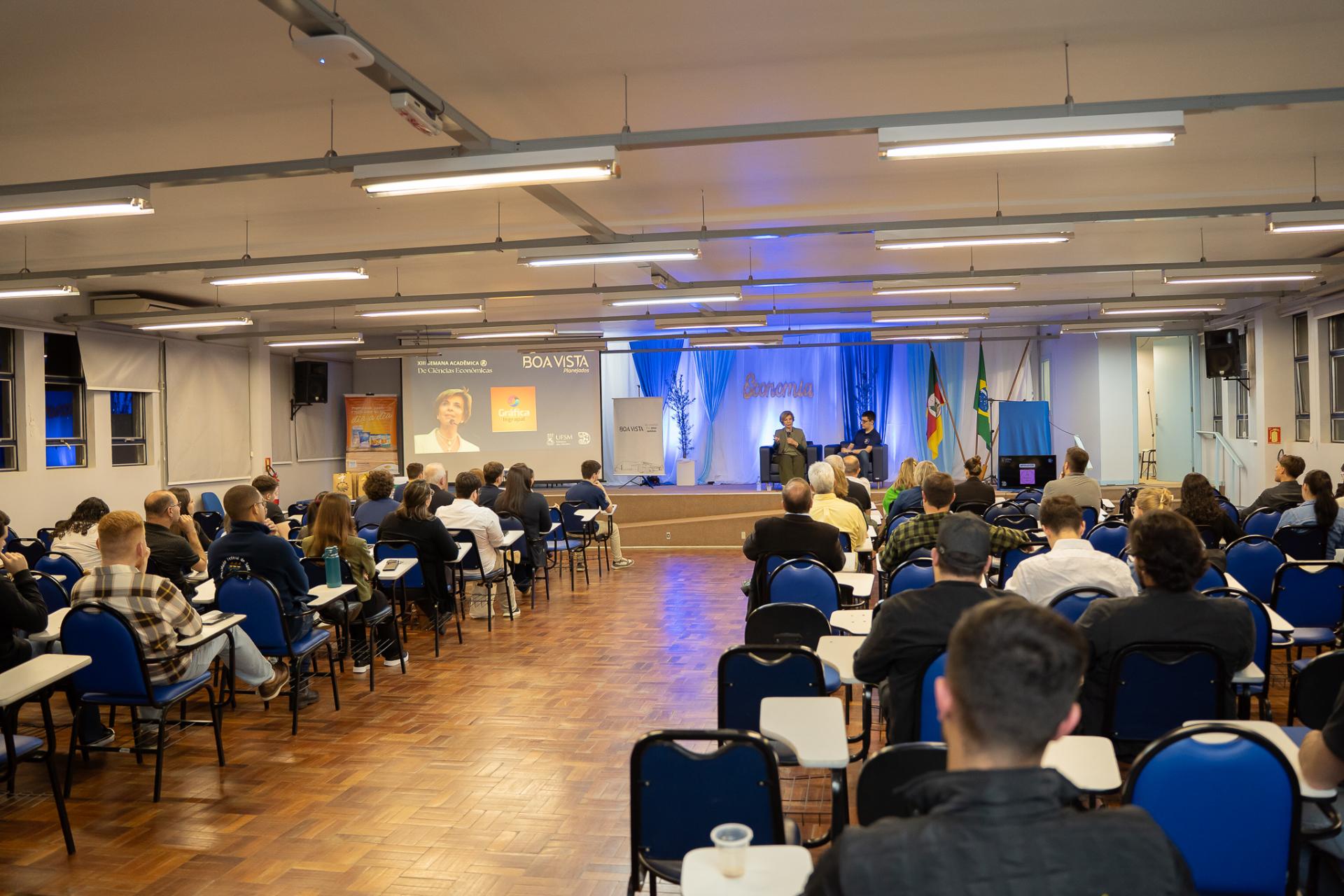 Foto colorida horizontal de auditório. Dos dois lados estão pessoas sentadas em cadeira alinhadas. Entre elas, um corredor. À frente, a ex-governadora Yeda Crusius e o apresentador do evento. 