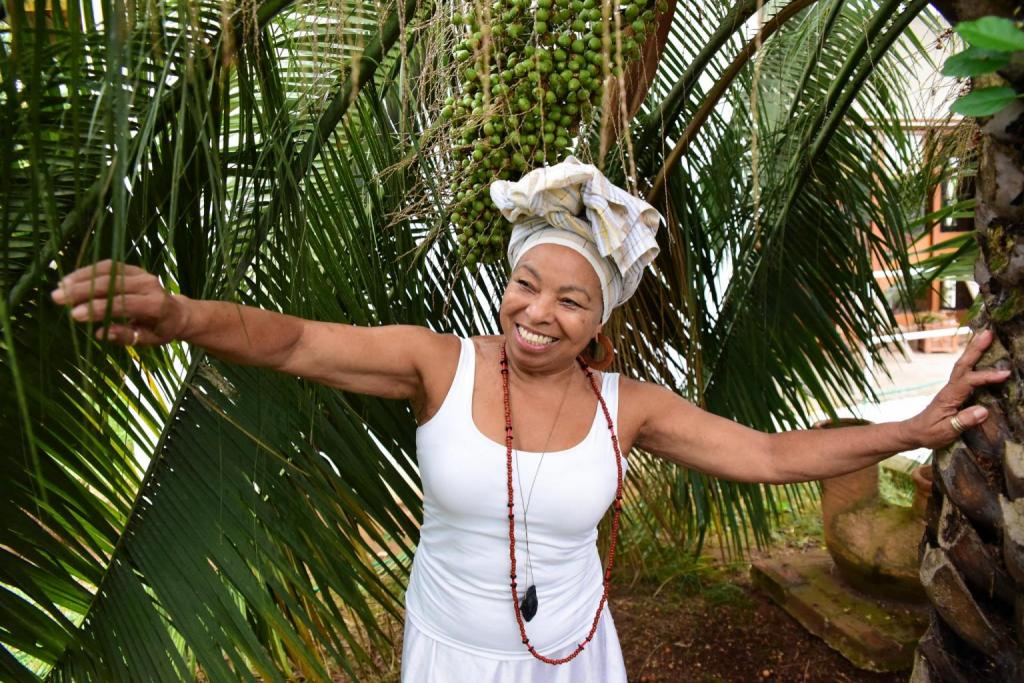 Foto colorida horizontal de mulher negra com roupa branca e turbante na cabeça. A mulher usa um colar, está de braços abertos e sorrindo. O local em que ela está tem folhas verdes largas