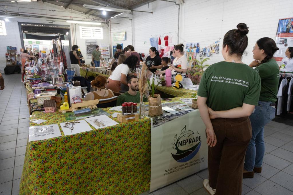 foto colorida horizontal de um estande no centro de um pavilhão. Há objetos dispostos sobre a bancada, um banner pendurado do Neprade e 3 pessoas de camiseta verde escuro ao redor