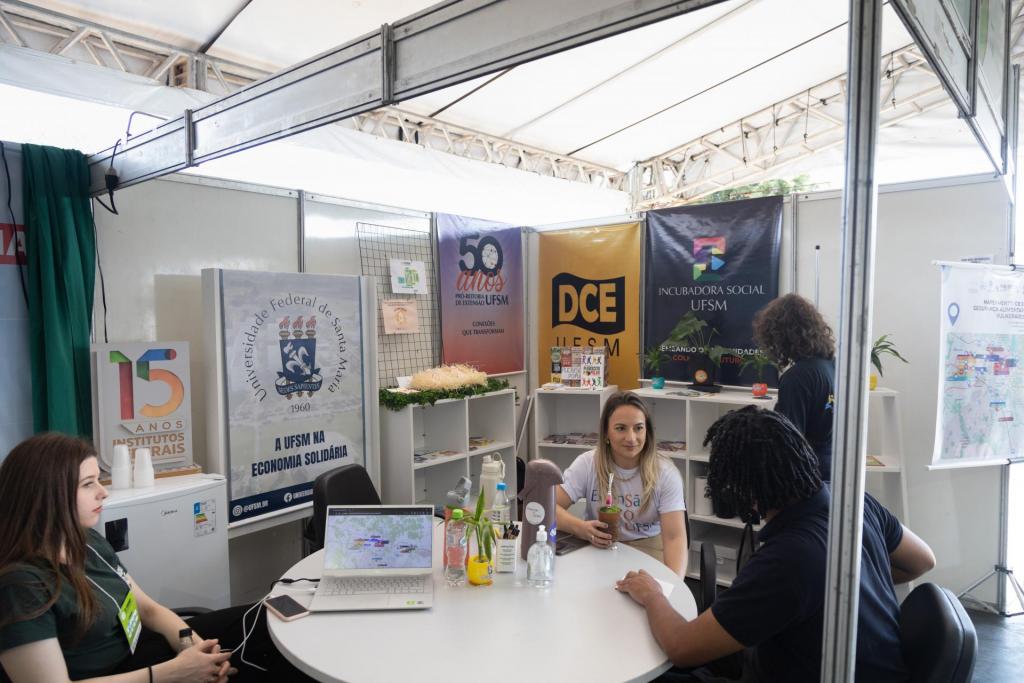 foto colorida horizontal de um estande interno com banners pendurados da UFSM, DCE, PRE e Incubadora Social. Há algumas pessoas sentadas ao redor de uma mesa