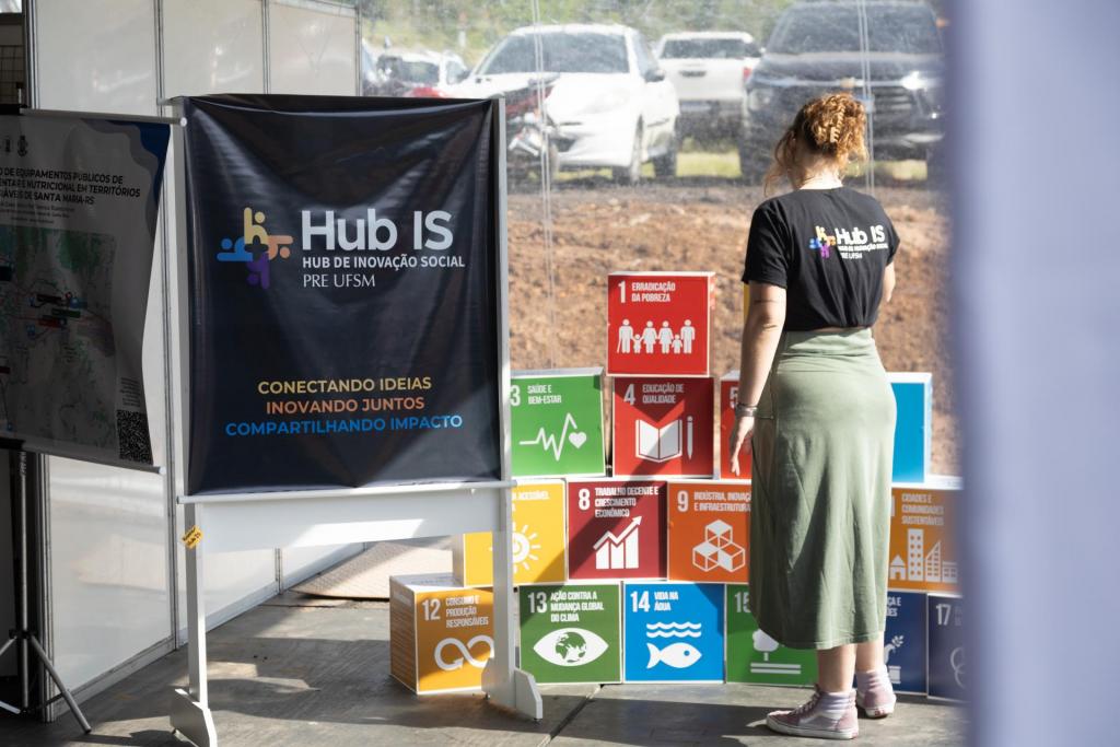 foto colorida horizontal de um espaço envidraçado com um banner preto do Hub IS, caixas coloridas mostrando cada um dos ODs e uma mulher de costas, olhando para as caixas