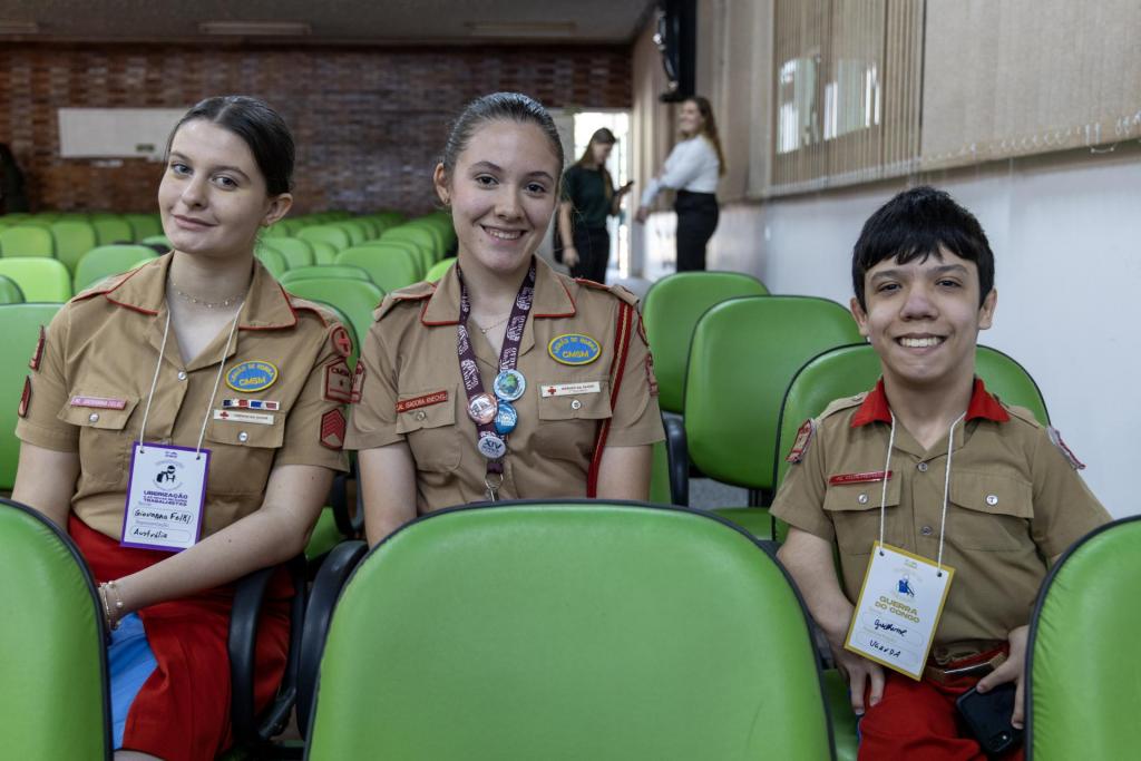 Foto colorida e horizontal. Três jovens estão sentados lado a lado em cadeiras verdes, sorrindo para a câmera. Todos vestem uniformes bege com detalhes vermelhos e crachás pendurados no pescoço. Ao fundo, o auditório está quase vazio, com fileiras de cadeiras verdes e duas pessoas de pé próximas à parede, em conversa.