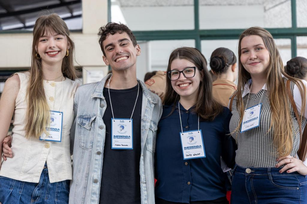 Foto colorida e horizontal. Três mulheres e um homem estão lado a lado, sorrindo para a câmera. Da esquerda para a direita: uma mulher de cabelo loiro veste blusa clara sem mangas; um homem de cabelo curto veste camiseta preta e jaqueta jeans; uma mulher de cabelo castanho e óculos veste camisa azul; e outra mulher de cabelo longo veste blusa xadrez com camisa branca por baixo. Todos usam crachás azuis do grupo “Imprensa Internacional”. Ao fundo, há pessoas de uniforme bege conversando em um ambiente interno com janelas amplas.