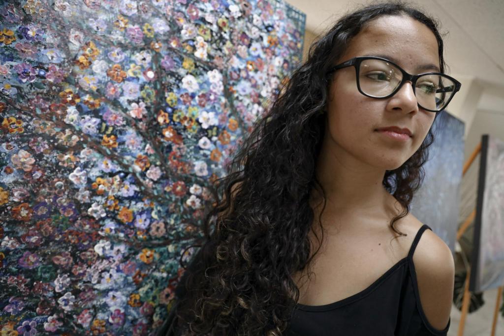 Foto colorida horizontal de Francine. A imagem traz o rosto da estudante, que está posicionada de forma inclinada à frente de um painel colorido da Biblioteca. No painel, uma árvore colorida