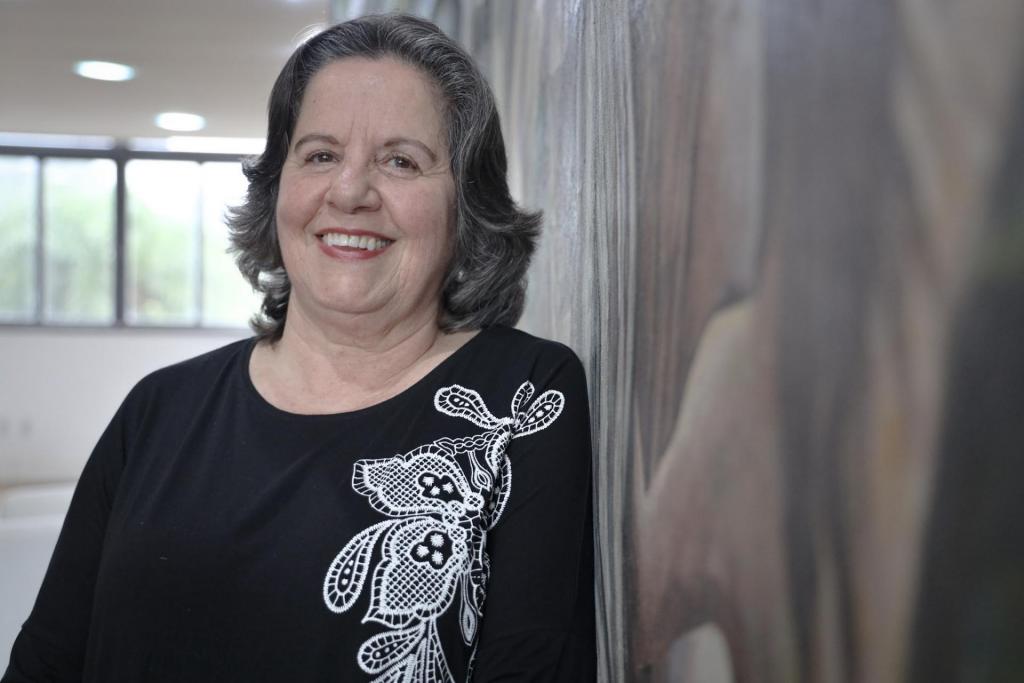 Foto colorida horizontal da Tânia na altura do peito. Ela está à erqueda, com cabelo solo e uma blusa preta com flores brancas. Tânia sorri para a câmera e está ao lado do painel de Eduardo Trevisan no Salão Imembuí