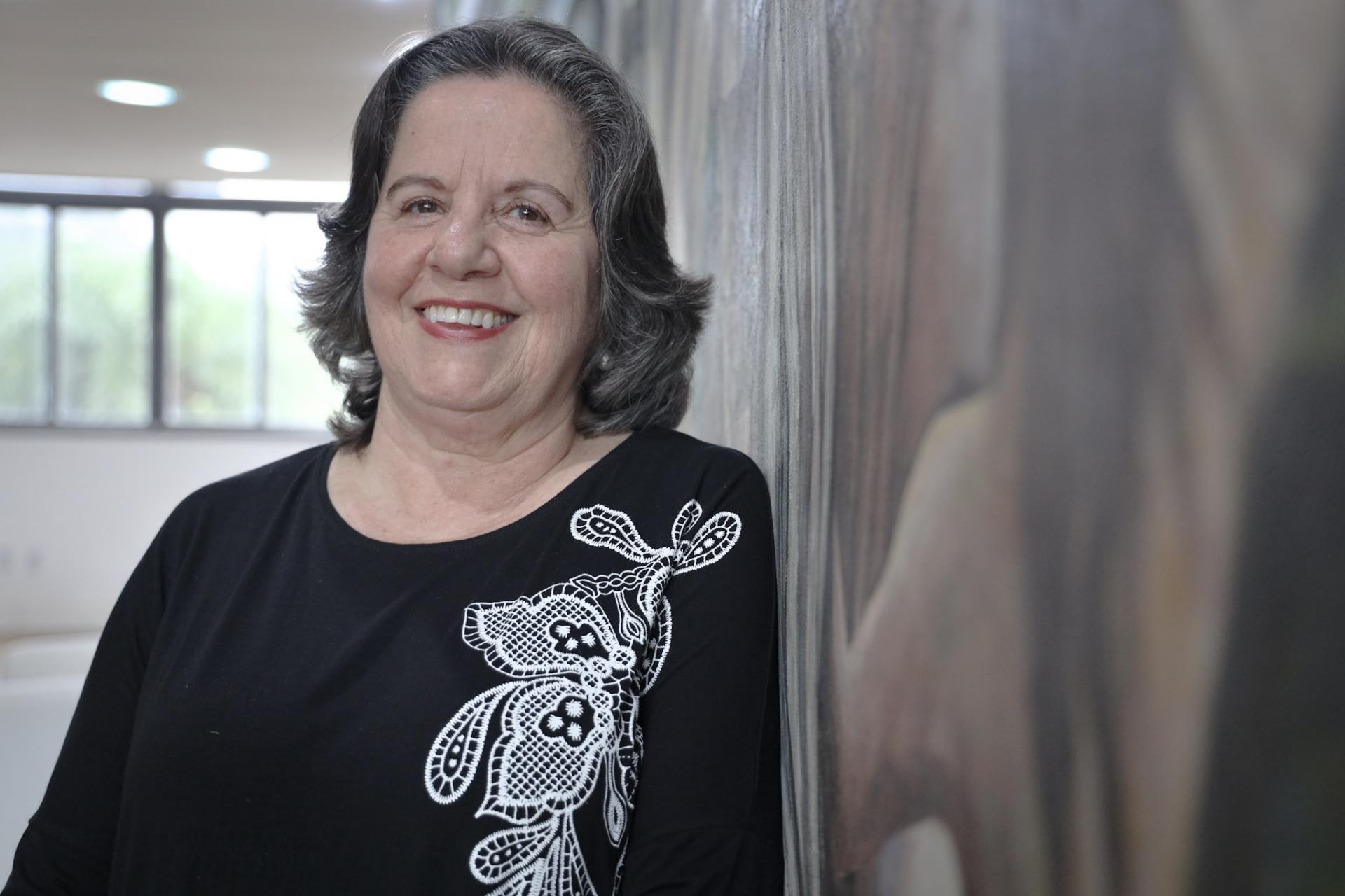Foto colorida horizontal da Tânia na altura do peito. Ela está à erqueda, com cabelo solo e uma blusa preta com flores brancas. Tânia sorri para a câmera e está ao lado do painel de Eduardo Trevisan no Salão Imembuí