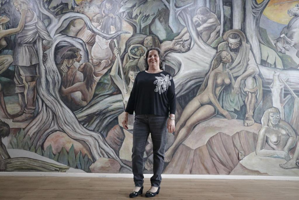 Foto colorida horizontal de Tânia à frente do painel "Imembuí" de Eduardo Trevisan. Tânia está ao centro, com cabelo solto, uma blusa preta, uma calça preta e um sapato de saltos. Atrás dela, o painel retrata a lenda de Imembuí