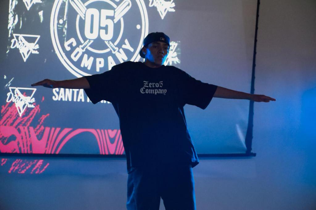 foto colorida horizontal de um rapaz com os braços abertos num palco semiescuro, com telão ao fundo