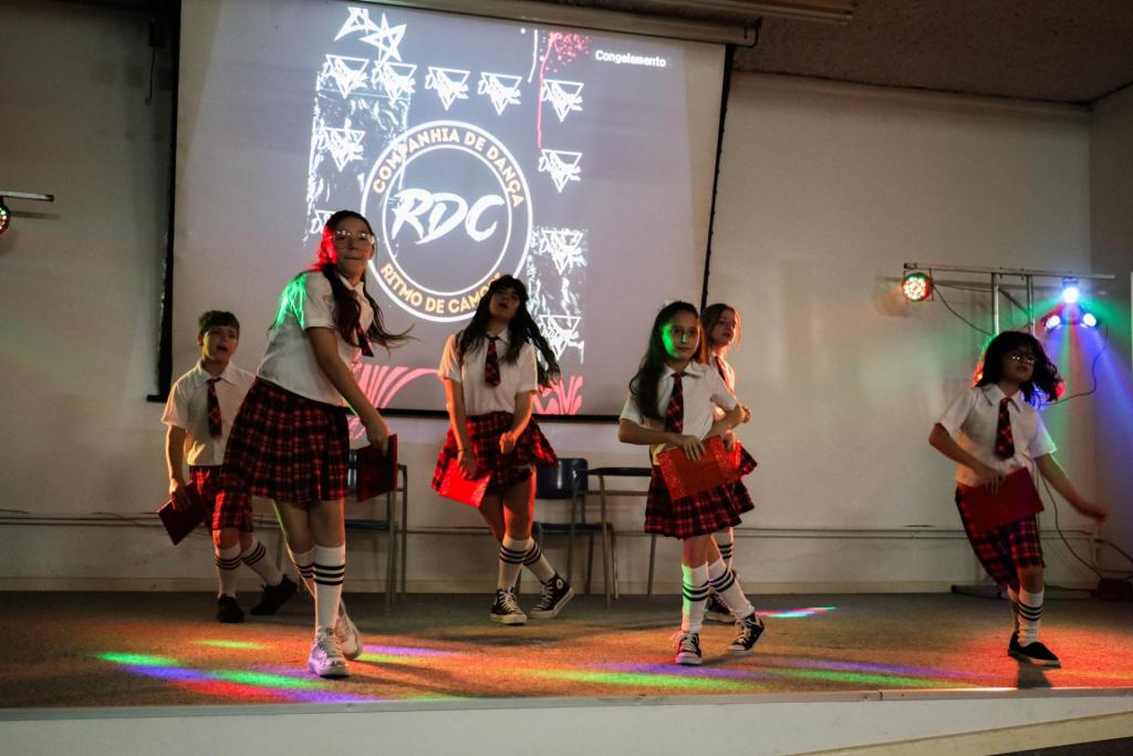 foto colorida horizontal de meninas danãndo num palco. Elas usam camias branca, gravata e saia rodada xadrez em tom vermelho