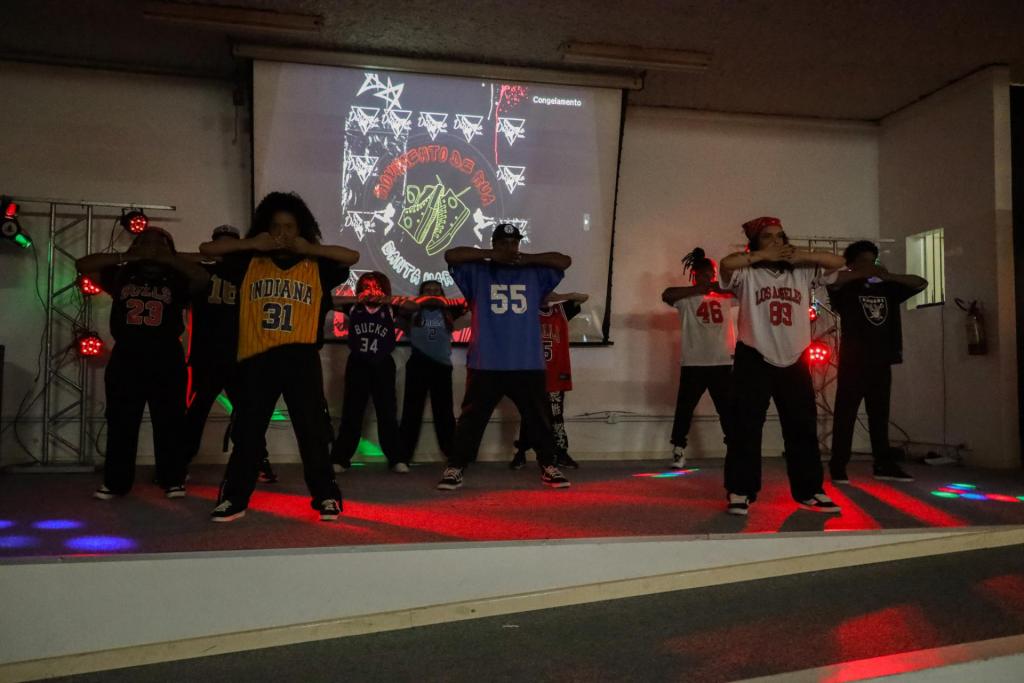 foto colorida horizontal de um grupo de jovens dançando num palco, de calça escura e camisetas de basquete com números na frente
