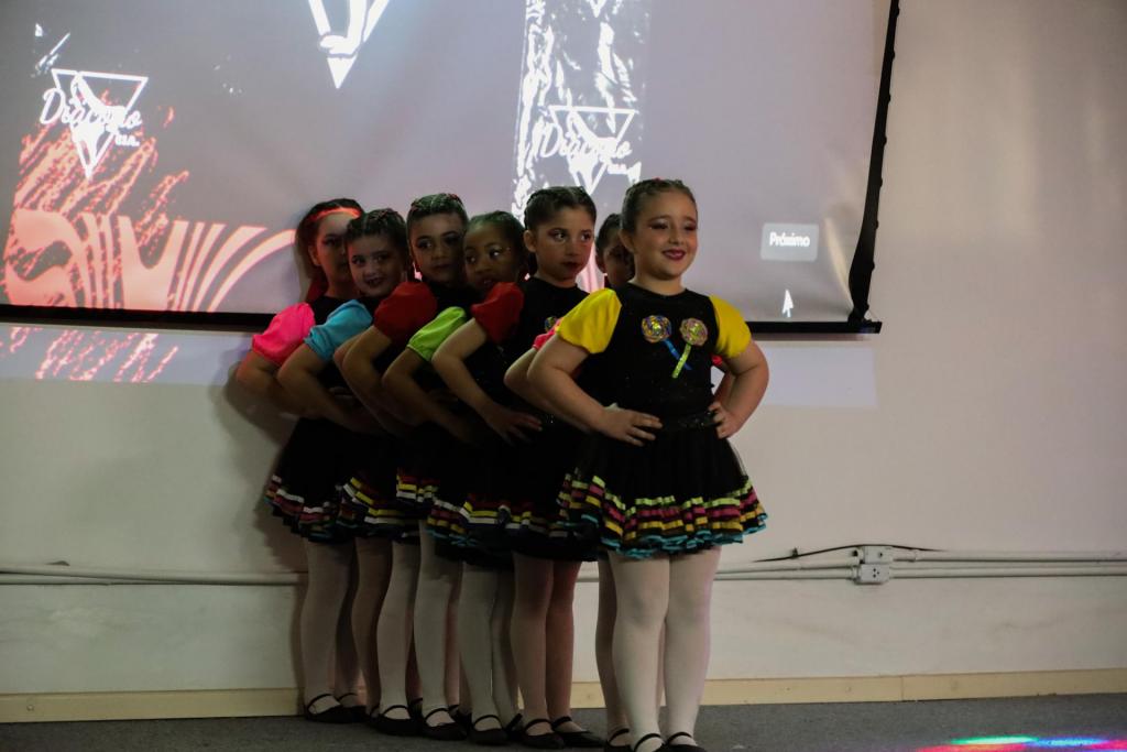 foto colorida horizontal de crianças, meninas, dançando num palco, de vestidos coloridos. Elas estão em fila e com as mãos na cintura