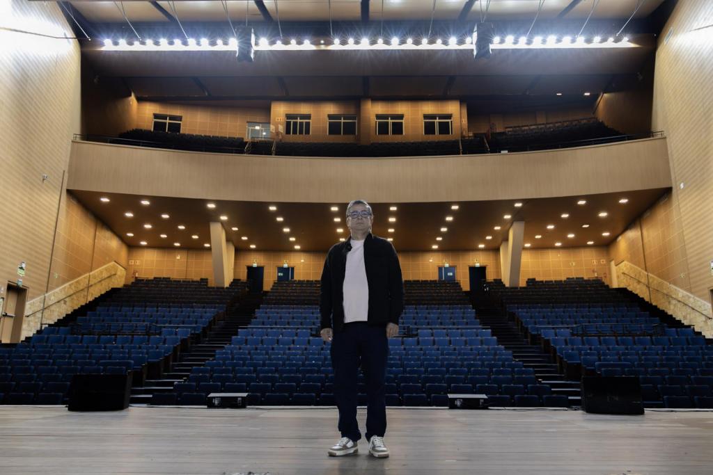 Foto colorida horizontal de Felipe, centralido à frente do palco e da plateia do Centro de Convenções. A foto mostra um homem bem ao centro e detalhes do lugar, como o palco, a plateia e o mezanino